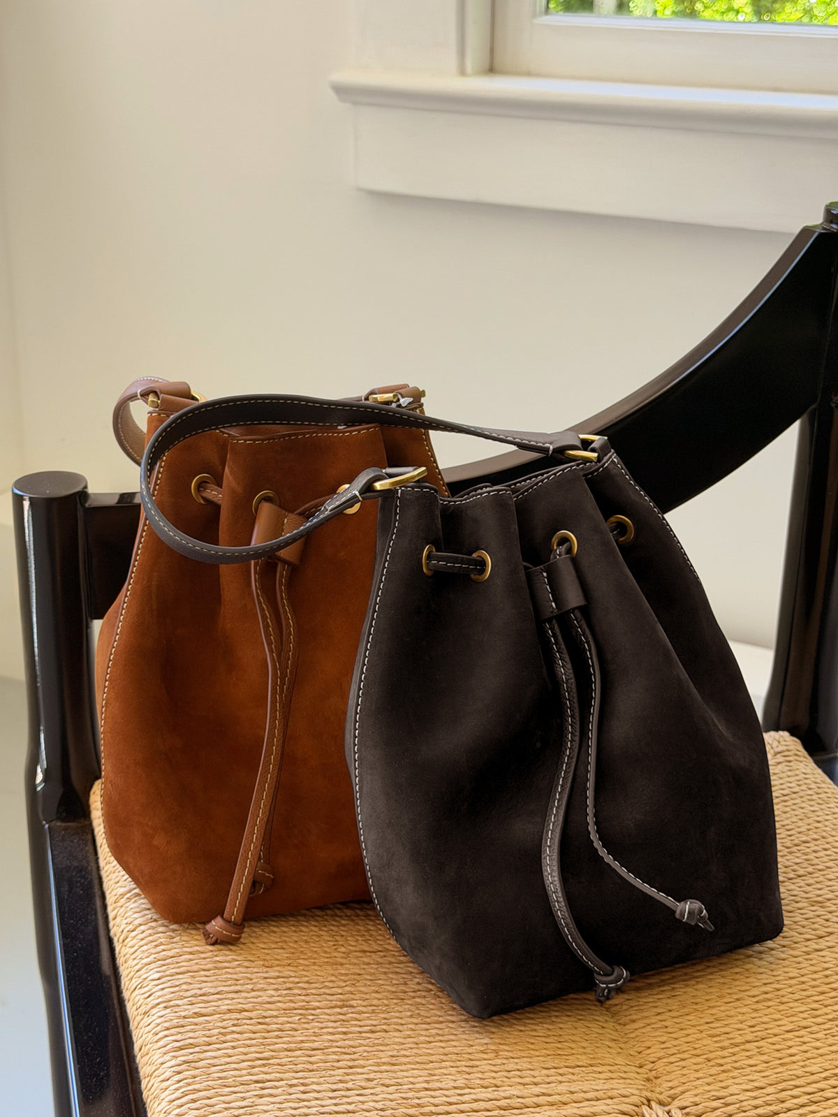 Two suede bags in rich brown and black tones with drawstring closures sit on a woven seat, showcasing The Newbury Suede Bucket Bag by J.McLaughlin’s timeless style and craftsmanship.