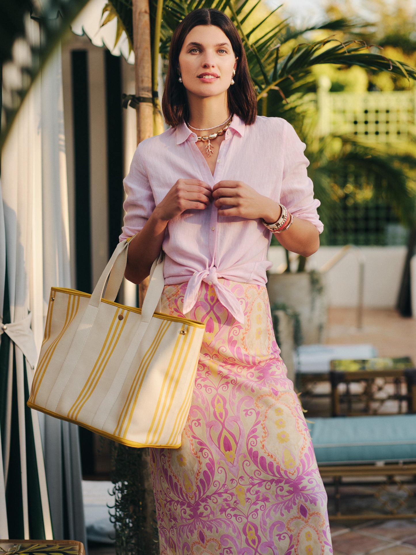 Woman standing outdoors wearing a pink blouse and patterned skirt, carrying the Alma Canvas Tote in Stripe by J.McLaughlin, showcasing effortlessly stylish bags and enduring designs.