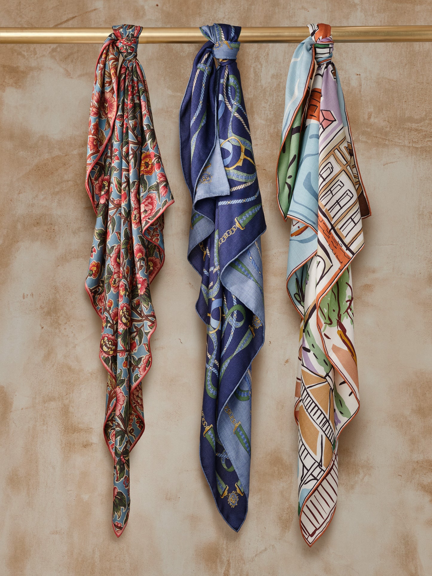 Three elegant Vanessa Scarf in Village Stroll Border designs from J.McLaughlin hanging on a wooden rod, showcasing effortlessly stylish scarves with enduring designs and a legacy of timeless fashion. A destination for defining style. Life, Well-Lived.