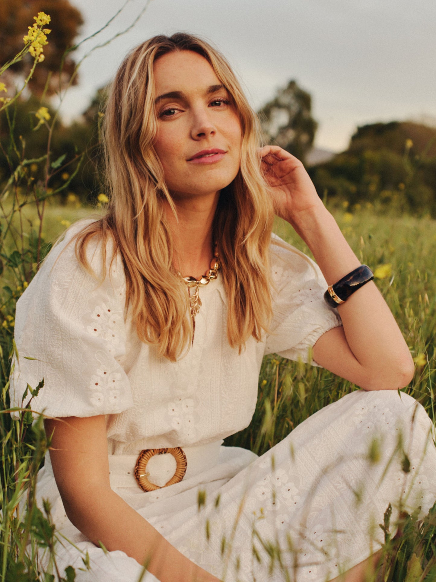 Woman seated in a field wearing a white eyelet dress and a Resin Hinge Bangle on her wrist, showcasing J.McLaughlin's effortlessly stylish, enduring designs and timeless fashion.