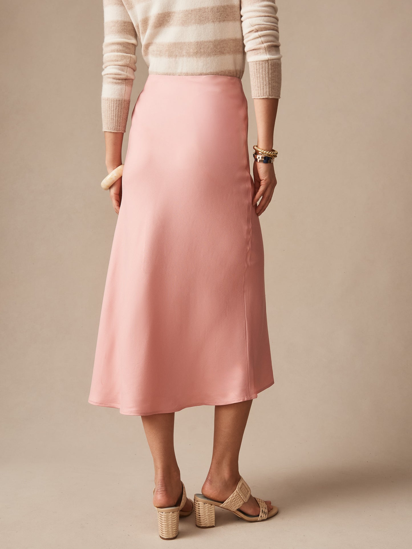 A back view of a woman wearing the Adelyn Skirt by J.McLaughlin in a soft pink shade paired with a striped sweater and heeled sandals, showcasing effortlessly stylish and enduring designs from a destination for defining style. Life, Well-Lived.