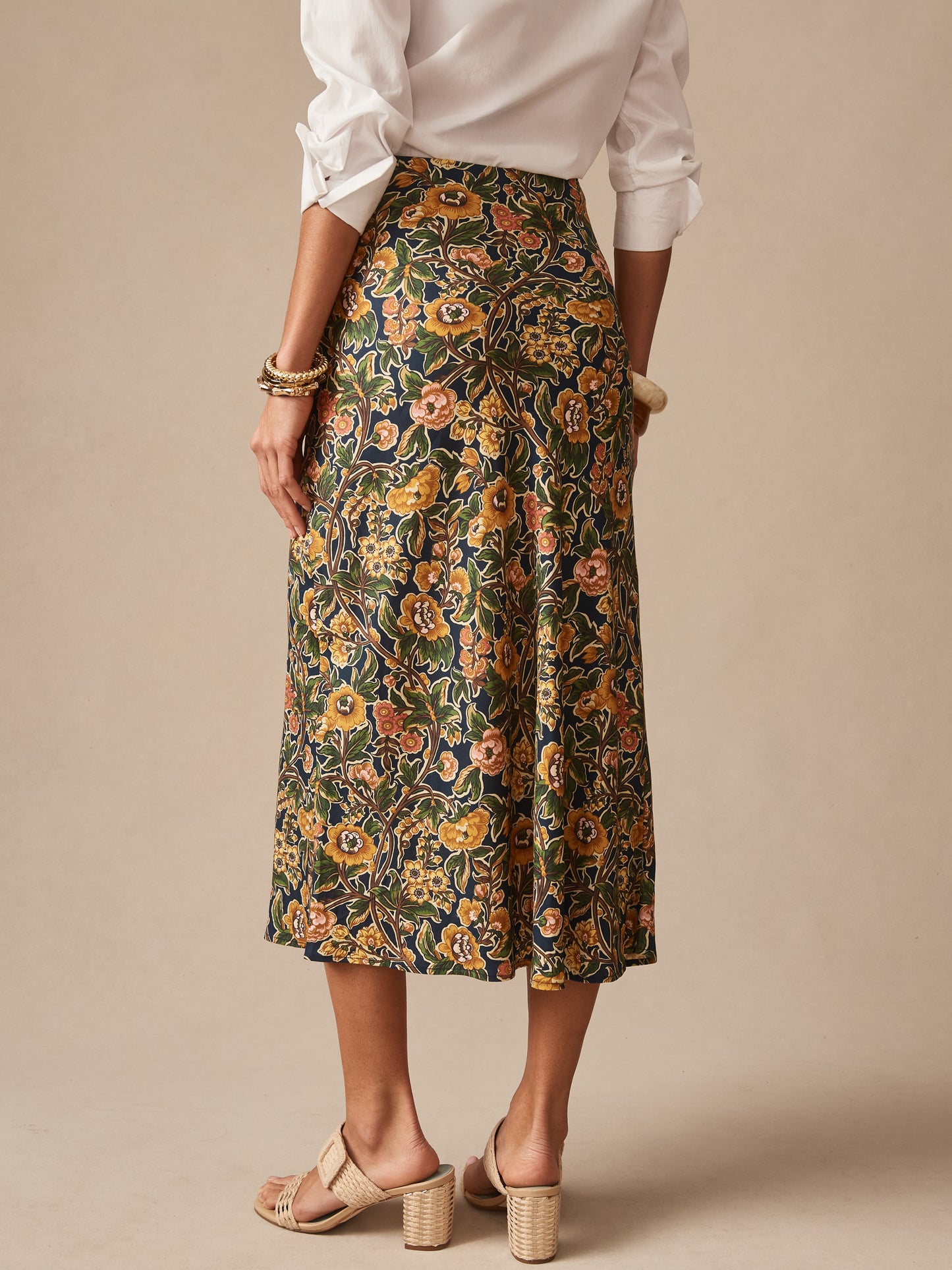 A woman models the Adelyn Skirt in Bucolic Blooms Bias by J.McLaughlin, featuring floral patterns in warm tones, paired with a white blouse and neutral heeled sandals, reflecting effortlessly stylish, enduring designs and a legacy of timeless fashion. A destination for defining style. Life, Well-Lived.