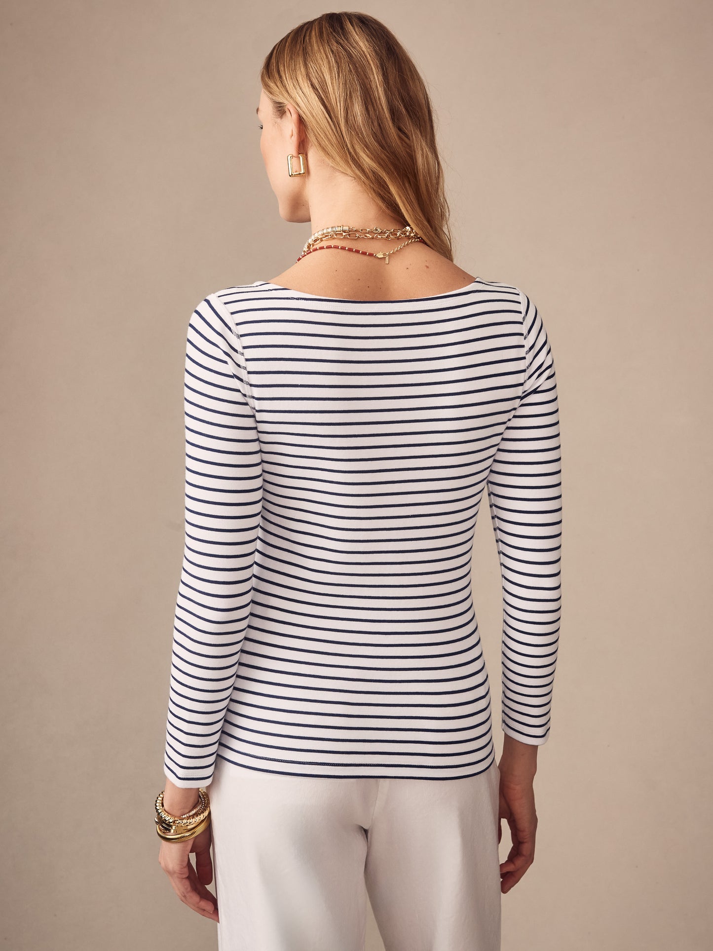 Woman wearing a Goldie Tee in Stripe by J.McLaughlin, featuring navy and white horizontal stripes and long sleeves, styled with gold jewelry and white pants, showcasing effortlessly stylish, enduring designs that reflect innovative fabrics and a legacy of timeless fashion at a destination for defining style. Life, Well-Lived.