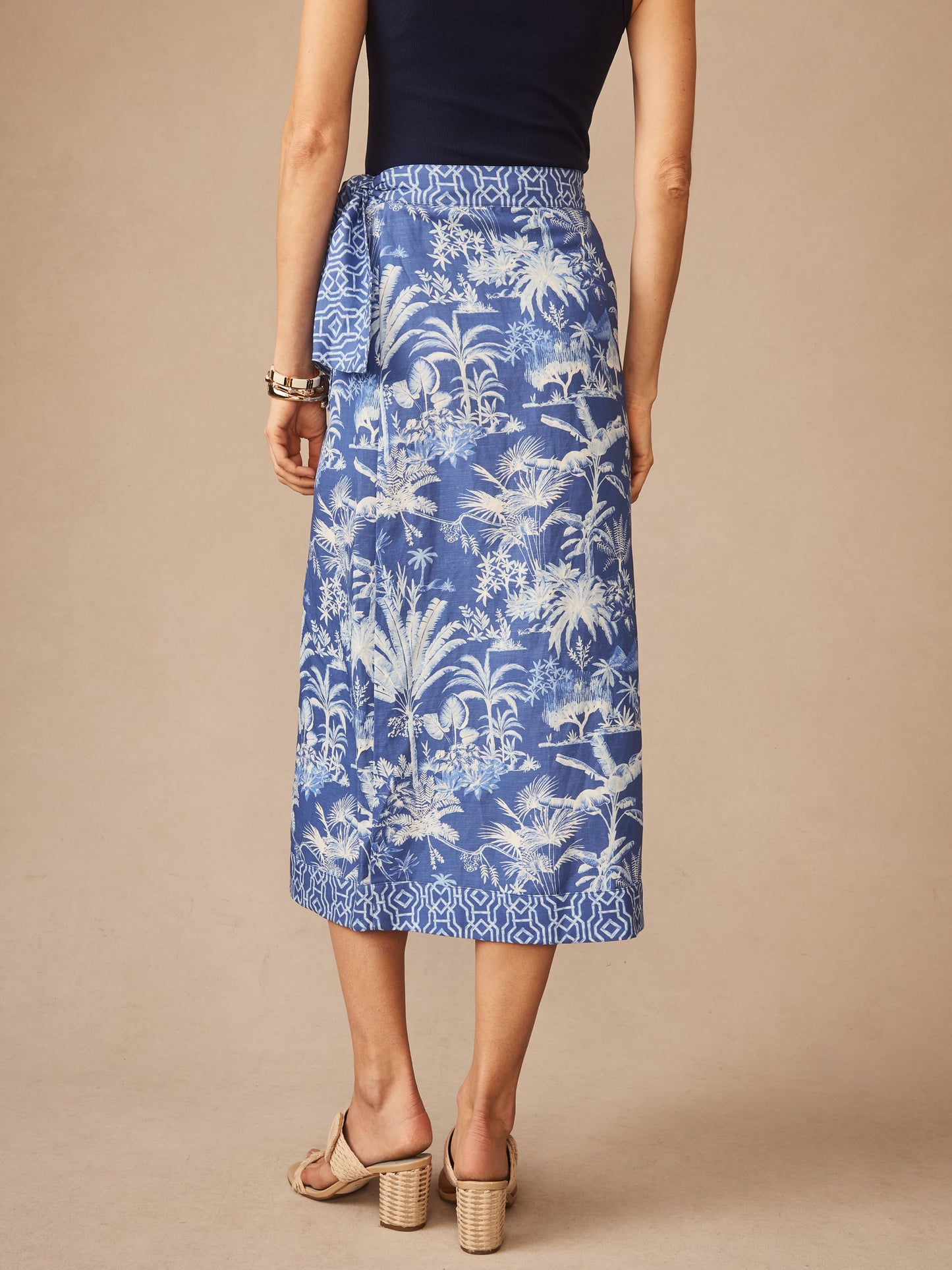 Back view of a woman wearing the Ardis Linen Blend Skirt in Garden Isle by J.McLaughlin, featuring a blue and white tropical print with a tied waist, paired with neutral heels, showcasing effortlessly stylish, enduring designs crafted from innovative fabrics, a destination for defining style. Life, Well-Lived.