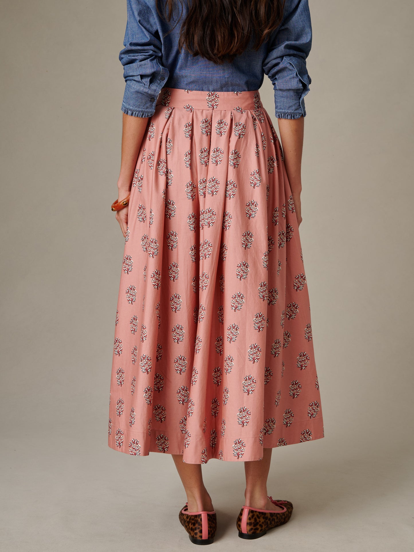 Back view of a woman wearing the Arabella Skirt in Heritage Bloom Cross by J.McLaughlin, featuring a flowing pink skirt with a floral pattern paired with a casual chambray shirt and leopard print flats.