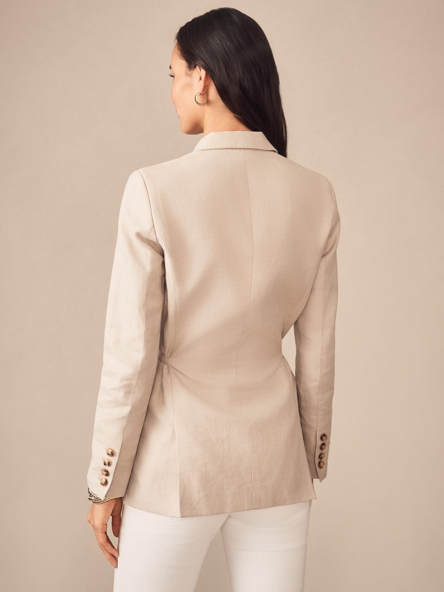 A woman wearing a sand-colored Biltmore Linen Blend Blazer from J.McLaughlin, showcasing the tailored back design with button details on the sleeves, embodying effortlessly stylish, enduring designs with innovative fabrics. A destination for defining style. Life, Well-Lived.