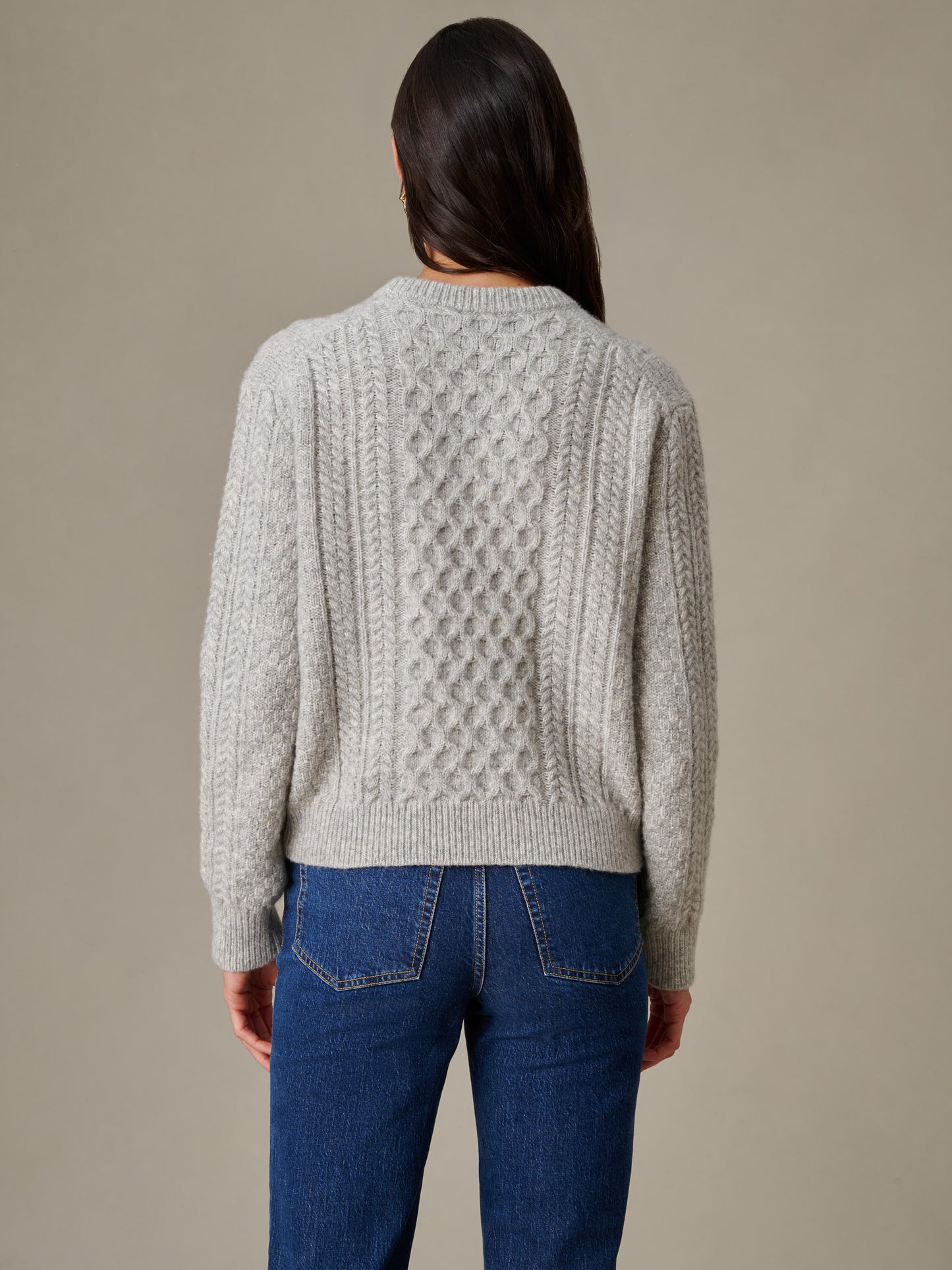 Woman wearing a light gray Brigette Sweater by J.McLaughlin featuring intricate cable knit design, paired with classic blue jeans, showcasing enduring style and craftsmanship.