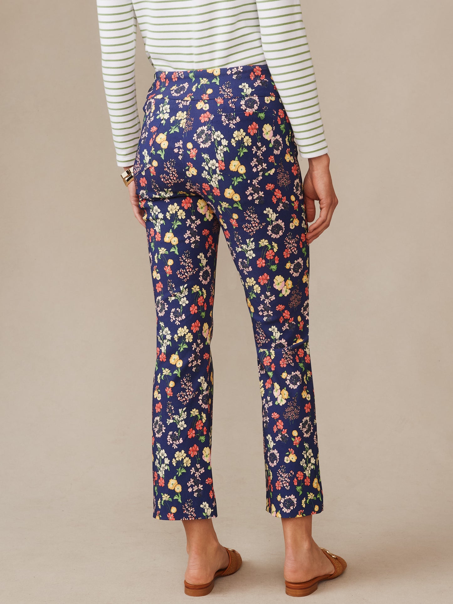 Woman seen from behind wearing Ivy Pants in Floral Medley by J.McLaughlin, featuring a navy base with colorful floral patterns, paired with a striped top and sandals; effortlessly stylish with enduring designs and innovative fabrics, reflecting a legacy of timeless fashion—a destination for defining style. Life, Well-Lived.
