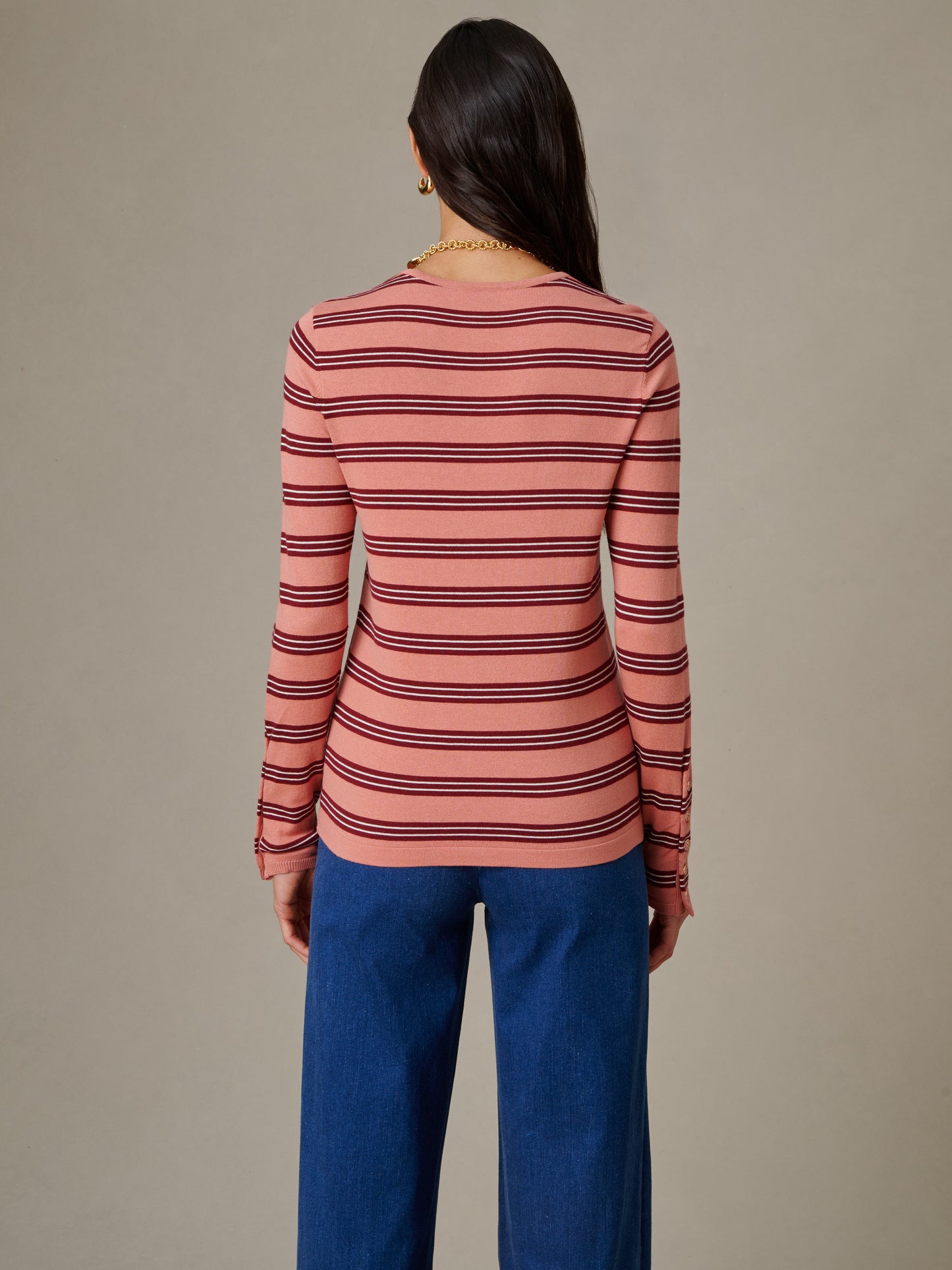 Woman seen from behind wearing a J.McLaughlin Jamey Sweater in Stripe with horizontal burgundy stripes on a muted pink background, paired with blue pants in a simple, timeless style.