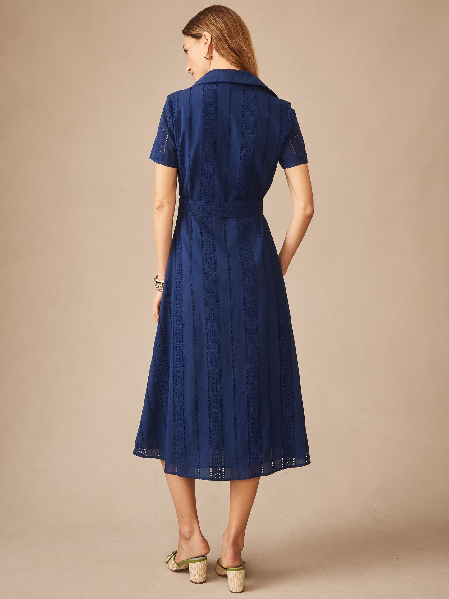 A woman models the Kalinda Dress in Eyelet by J.McLaughlin, featuring a navy blue, eyelet-patterned fabric with a classic collar and short sleeves, embodying effortlessly stylish, enduring designs and innovative fabrics in a legacy of timeless fashion. A destination for defining style. Life, Well-Lived.