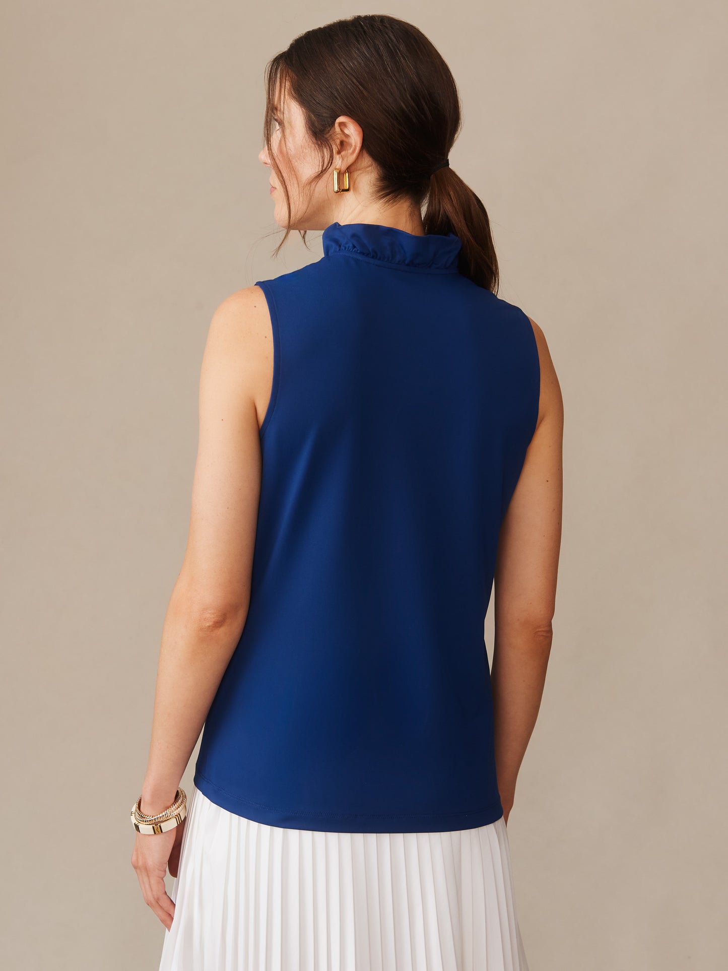 Woman wearing a navy Kate Sleeveless Ruffle Top by J.McLaughlin, featuring a ruffled high neck and sleeveless design, paired with a white pleated skirt, showcasing effortlessly stylish and enduring designs. A destination for defining style. Life, Well-Lived.