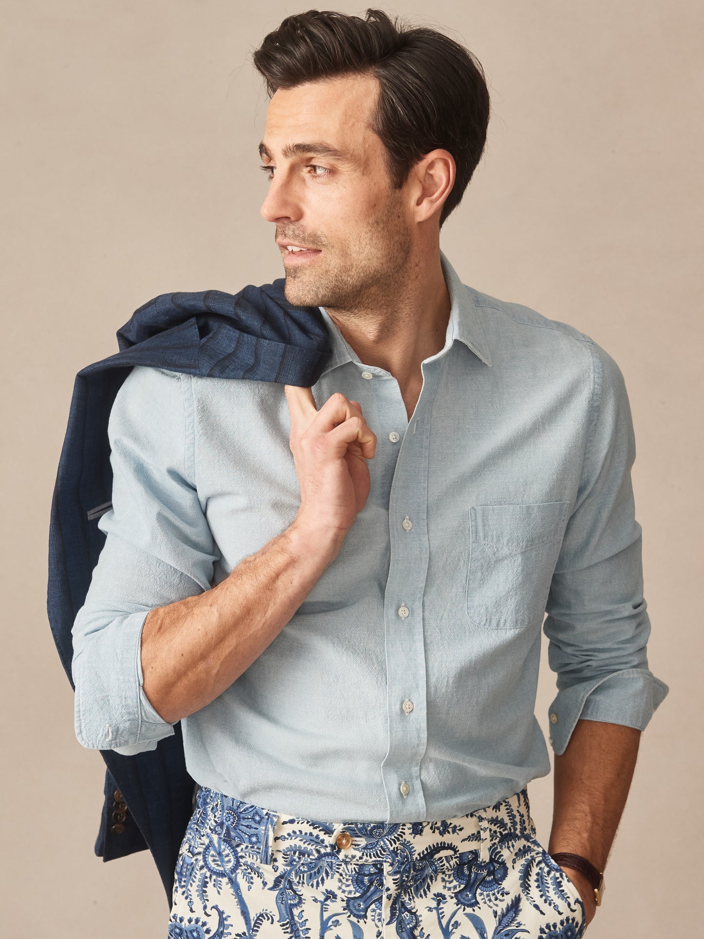 Man wearing a light blue Gramercy Cotton Chambray Shirt by J.McLaughlin, paired with patterned pants and holding a navy jacket over his shoulder, showcasing effortlessly stylish, enduring designs and innovative fabrics in a legacy of timeless fashion. A destination for defining style. Life, Well-Lived.