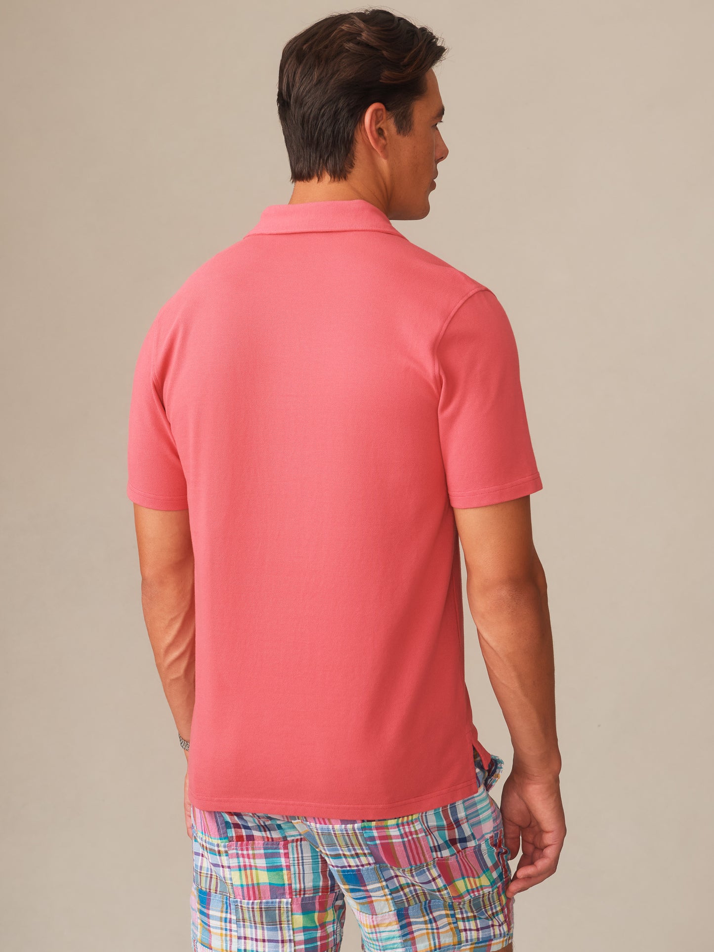 Man wearing a coral Mantle Pique Polo by J.McLaughlin paired with vibrant plaid shorts, showcasing effortlessly stylish, enduring designs with innovative fabrics in a legacy of timeless fashion. A destination for defining style. Life, Well-Lived.