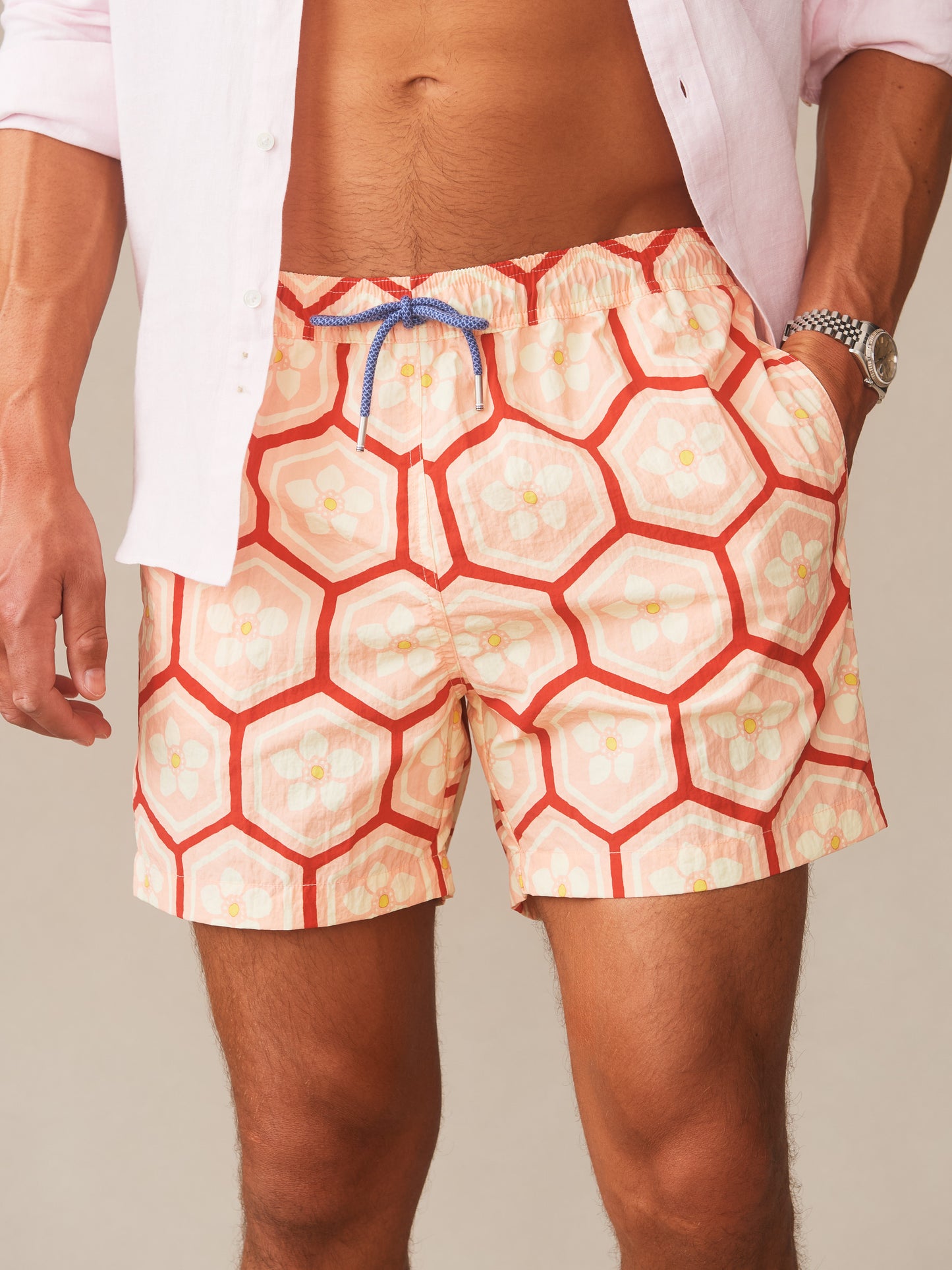 Man wearing Marina Swim Trunks in Daisy Postiano by J.McLaughlin, featuring a floral and geometric pattern in coral tones, paired with a light shirt and watch, showcasing effortlessly stylish swimwear and enduring designs from a destination for defining style. Life, Well-Lived.