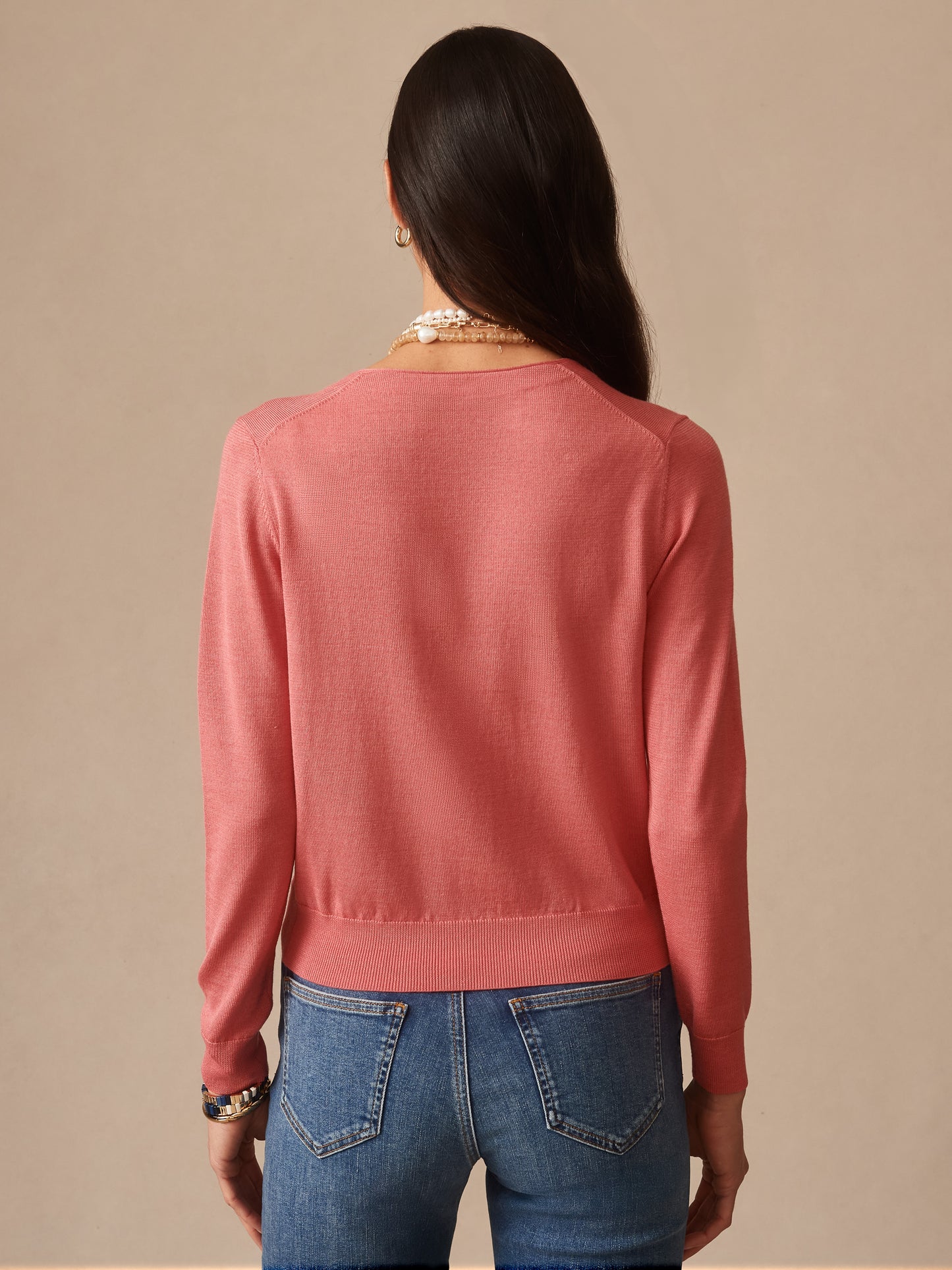 Woman wearing a soft coral Olivette Cardigan by J.McLaughlin, shown from the back with long sleeves and a fitted hem, paired with blue jeans.