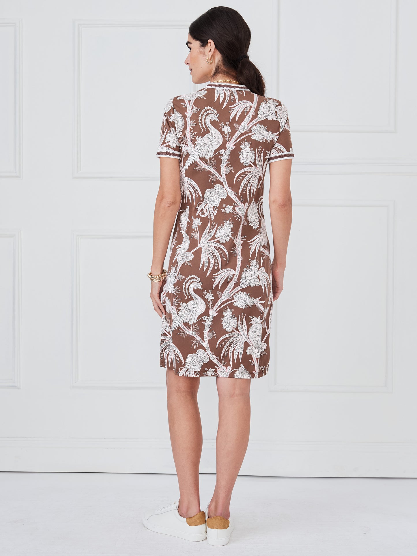 A woman wearing a J.McLaughlin Tesni Dress in Pomegrante Meadow with a brown base and white intricate bird and floral patterns, styled with casual white sneakers.