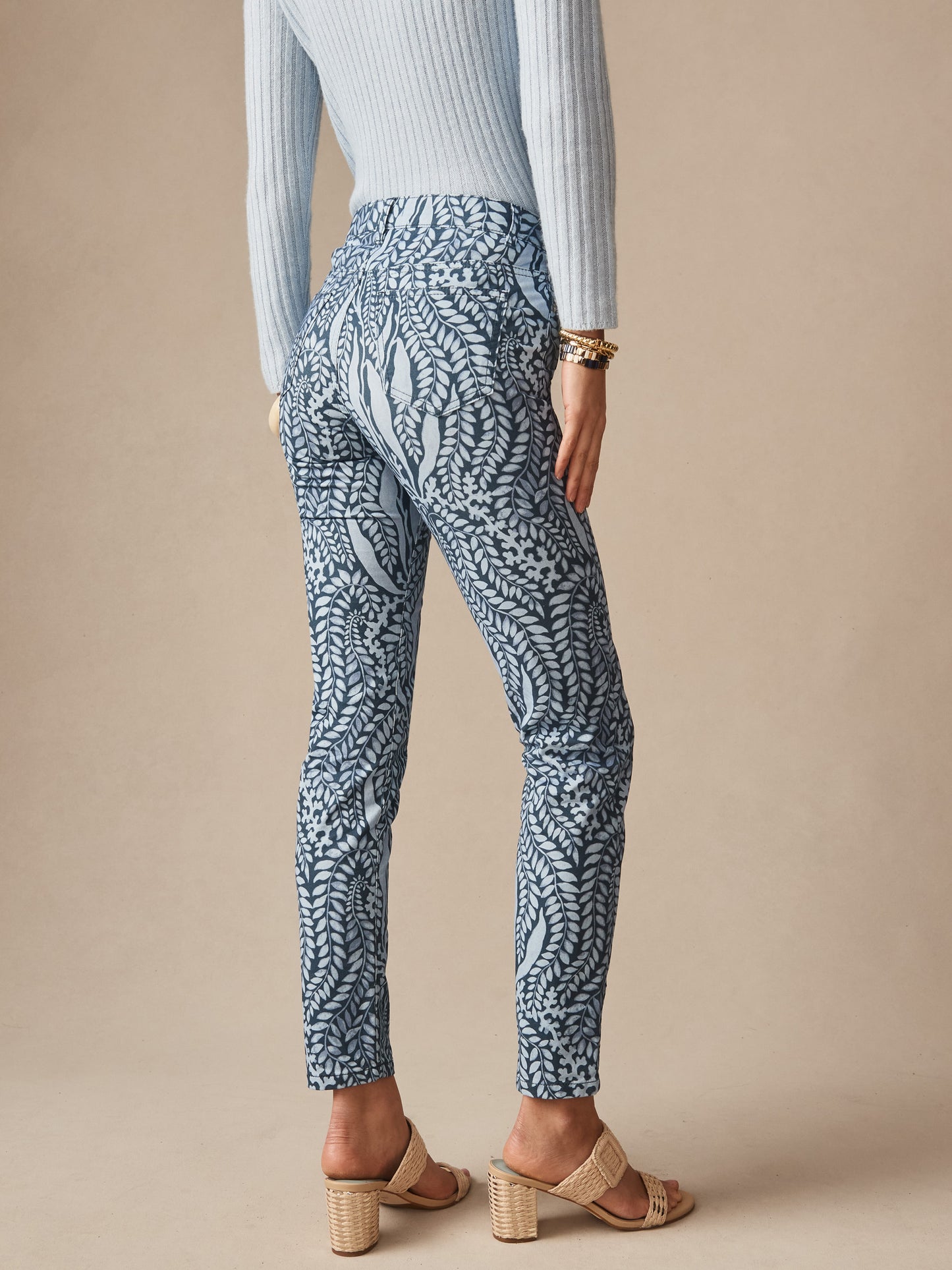 A model wearing J.McLaughlin Watson Cotton Cashmere Pants in Coral Current featuring a stylish blue leaf pattern, paired with neutral heels and a light ribbed sweater, showcasing effortlessly stylish, enduring designs with innovative fabrics and a legacy of timeless fashion. A destination for defining style. Life, Well-Lived.