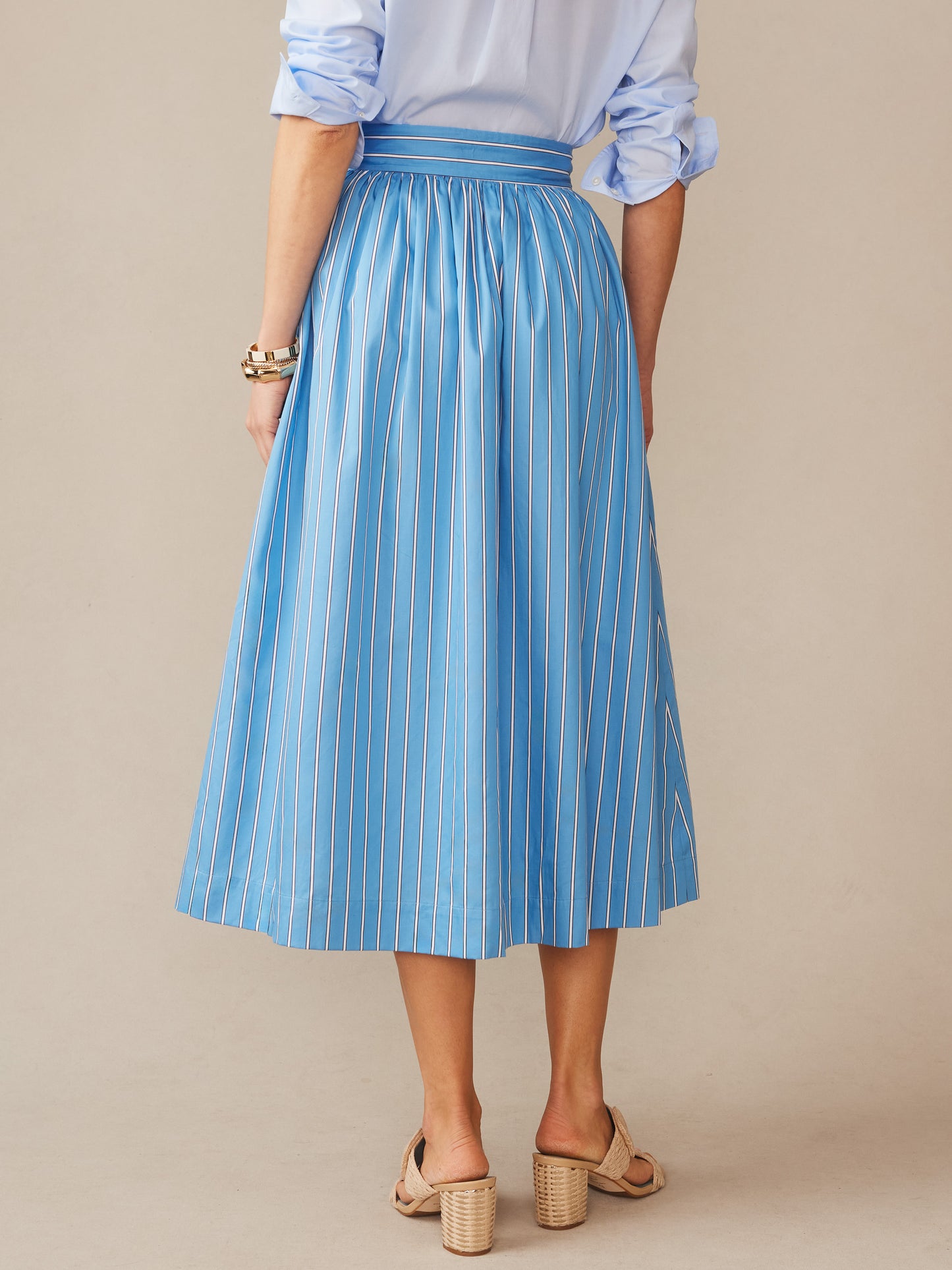A woman wearing the Winnie Skirt in Stripe by J.McLaughlin, featuring a flowing blue and white striped pattern paired with a rolled-sleeve shirt and beige heels, effortlessly stylish with enduring designs and innovative fabrics, embodying a legacy of timeless fashion— a destination for defining style. Life, Well-Lived.