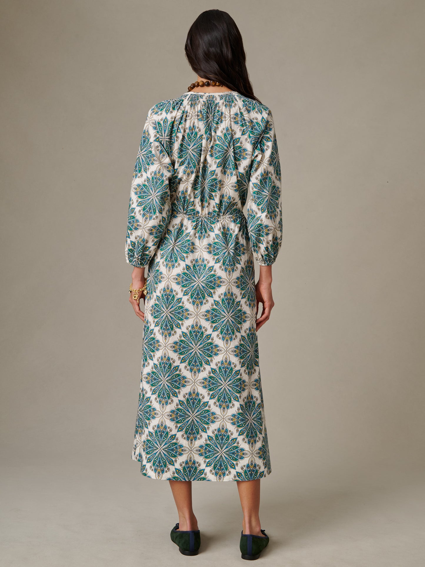 Back view of a woman wearing a long-sleeve, patterned dress with blue and green geometric floral prints, styled with simple dark shoes and jewelry, showcasing the Winslet Dress in Neo Radiant by J.McLaughlin.