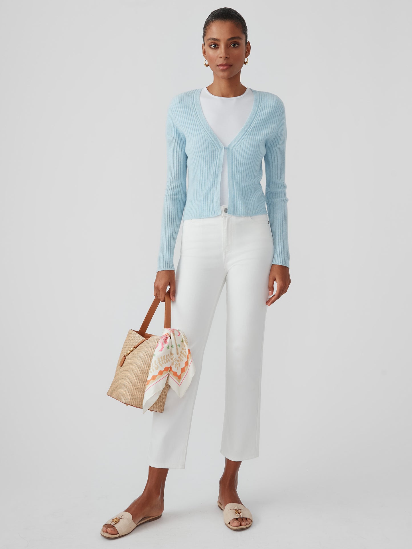Model wearing an effortlessly stylish baby blue Imogen Cashmere Cardigan by J.McLaughlin, paired with white pants and a woven bag, embodying enduring designs and timeless fashion.
