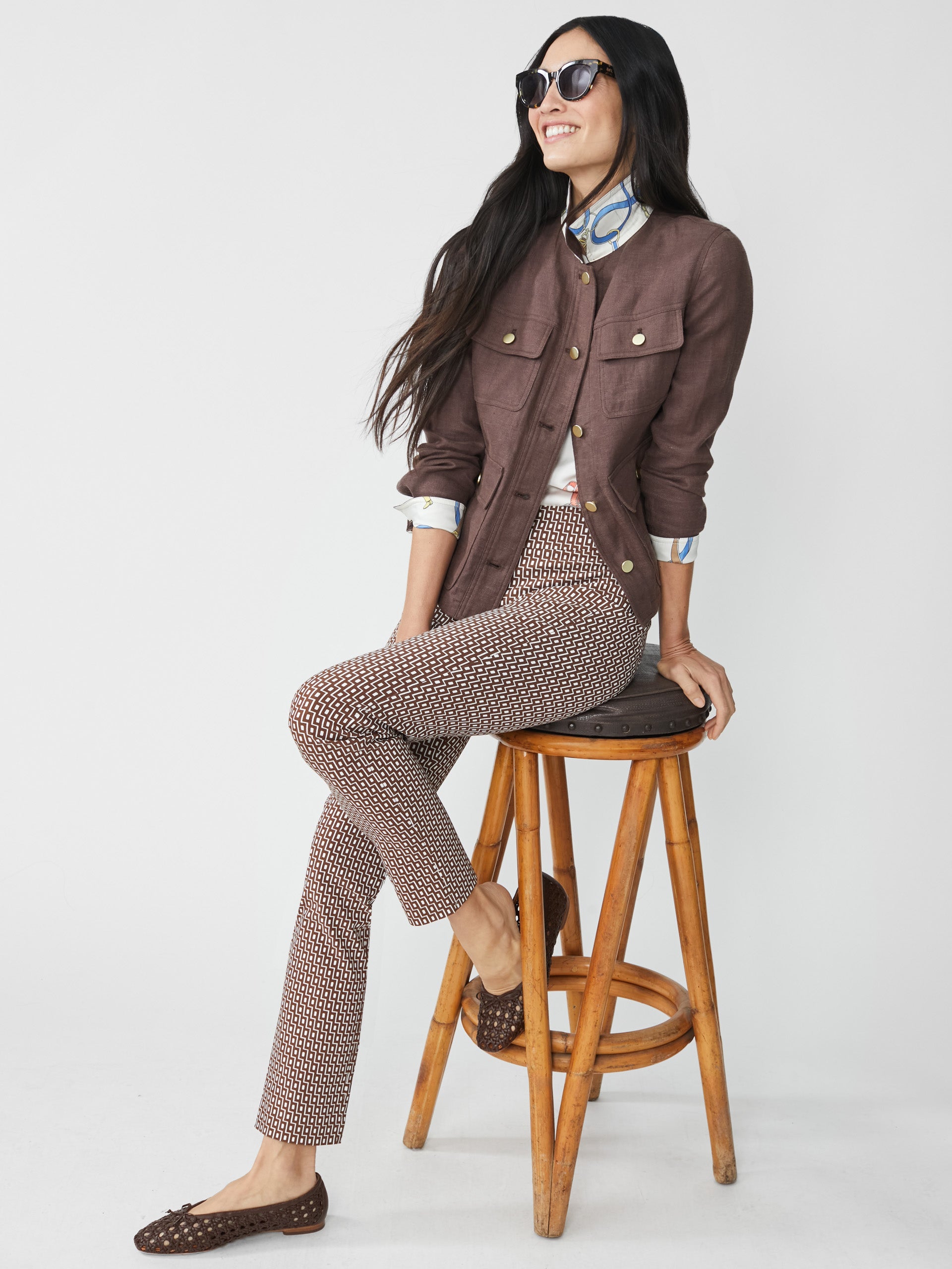 Woman seated on wooden stool wearing Masie Pants in Micro City Windows by J.McLaughlin, paired with a brown jacket and patterned blouse, showcasing effortlessly stylish, enduring designs and innovative fabrics. A destination for defining style. Life, Well-Lived.