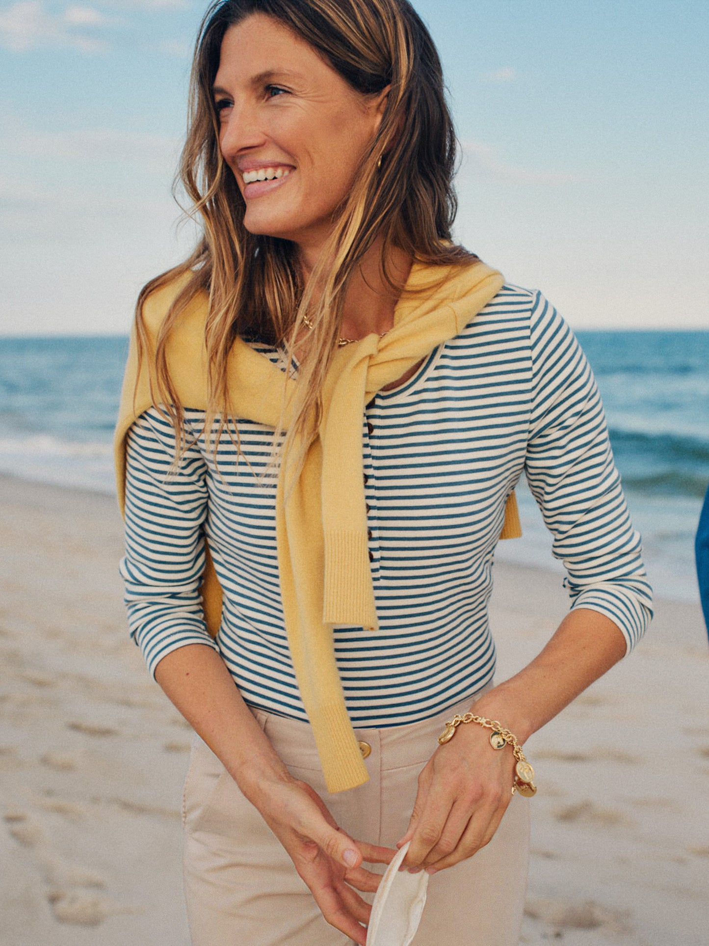 Woman smiling on a beach wearing a Sinclair Henley in Stripe by J.McLaughlin, styled with a yellow sweater draped over shoulders and beige pants, embodying effortless style.