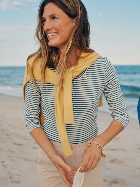 Woman smiling on a beach wearing a Sinclair Henley in Stripe by J.McLaughlin, styled with a yellow sweater draped over shoulders and beige pants, embodying effortless style.