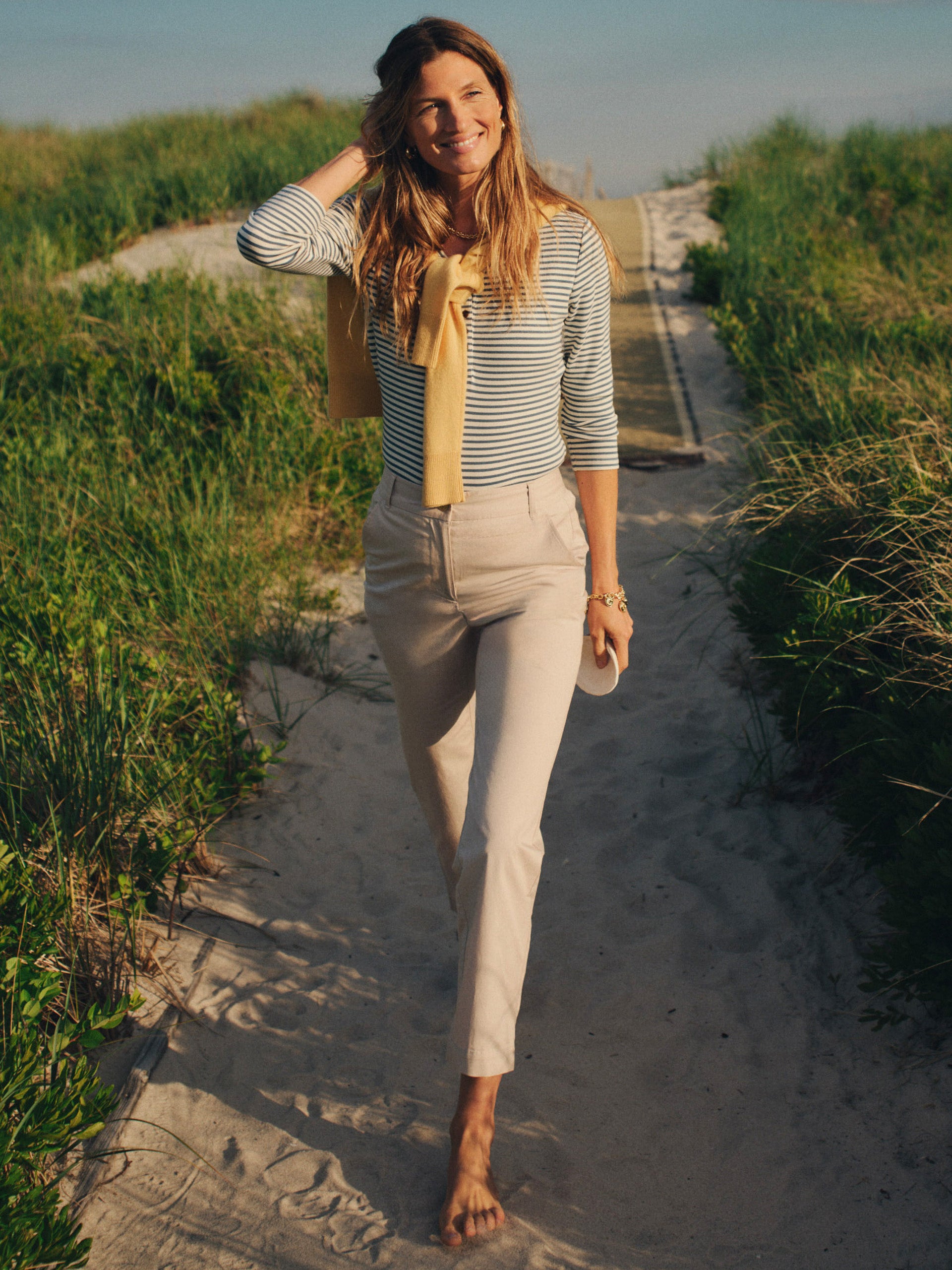Woman walking barefoot on a sandy path wearing Avery Cotton Chino Pants by J.McLaughlin, styled with a striped top and a yellow sweater draped over her shoulders, showcasing effortlessly stylish, enduring designs.