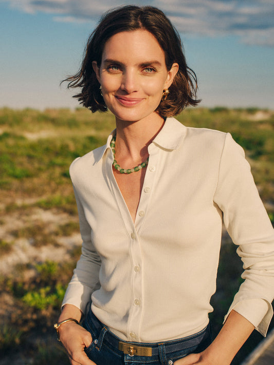 Woman wearing a cream Theodora Shirt by J.McLaughlin with a beaded necklace, standing outdoors in natural light embodying effortlessly stylish and enduring designs.