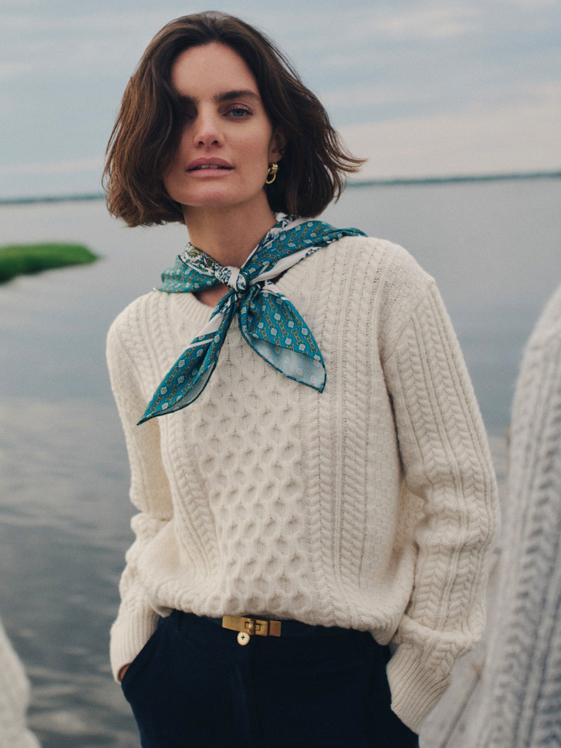 Woman wearing a cream cable-knit Brigette Sweater by J.McLaughlin paired with navy pants and a teal patterned scarf, showcasing timeless, effortlessly stylish fashion by the water.