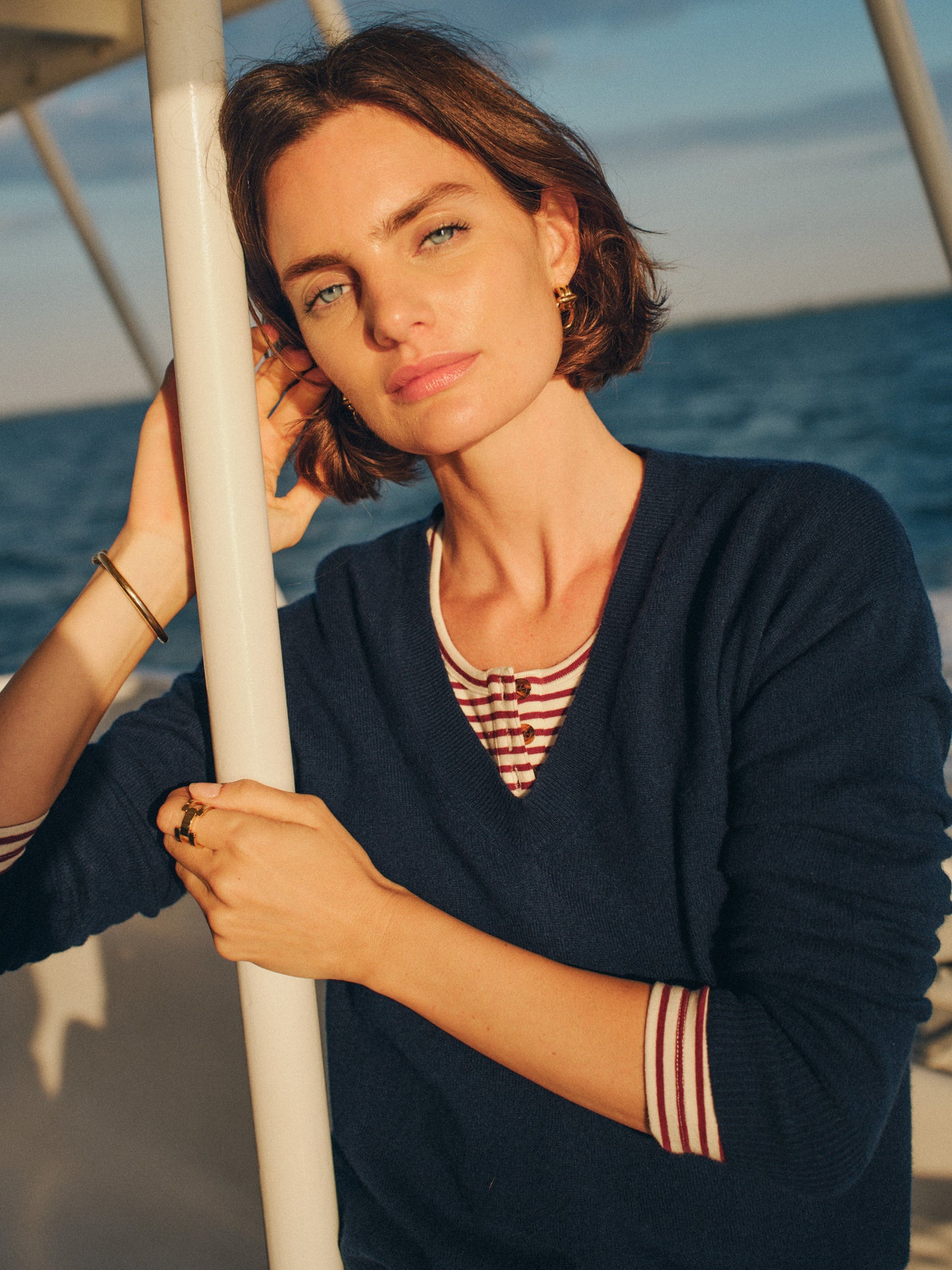 Woman wearing a navy Kerry Cashmere Sweater by J.McLaughlin, styled with a striped shirt underneath, posing outdoors near the ocean in warm sunlight.