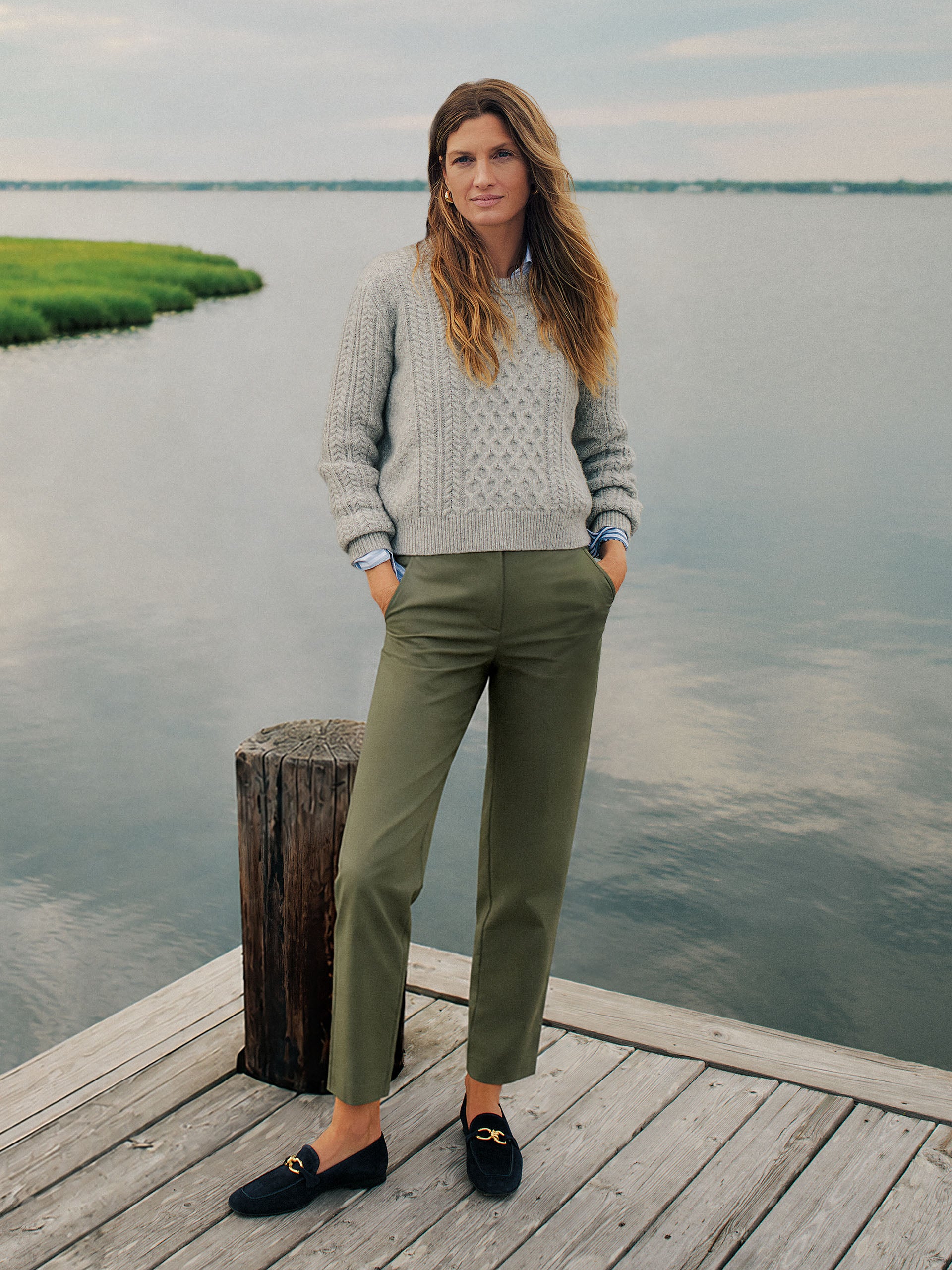 Woman standing on a dock wearing Avery Cotton Chino Pants by J.McLaughlin paired with a cozy gray sweater and black loafers, embodying effortlessly stylish, enduring designs.