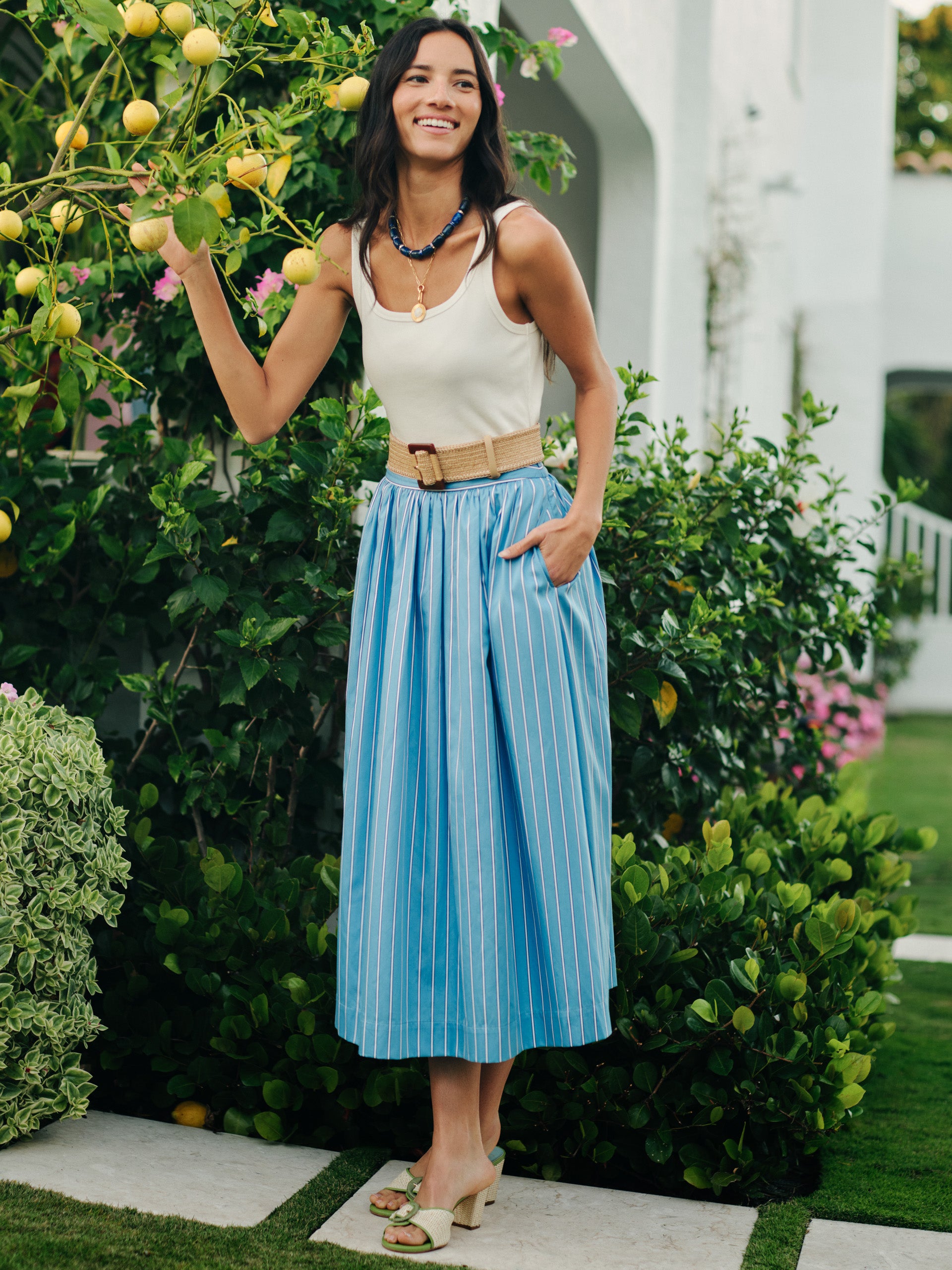 Woman wearing a white tank top with a blue striped Winnie Skirt in Stripe by J.McLaughlin, styled with natural accessories, showcasing effortlessly stylish, enduring designs in a garden setting.