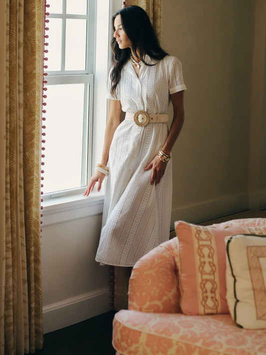 A woman wearing a Kalinda Dress in Eyelet by J.McLaughlin stands near a window in a softly lit room, showcasing effortless style and enduring design with a woven belt and delicate accessories, embodying a destination for defining style. Life, Well-Lived.