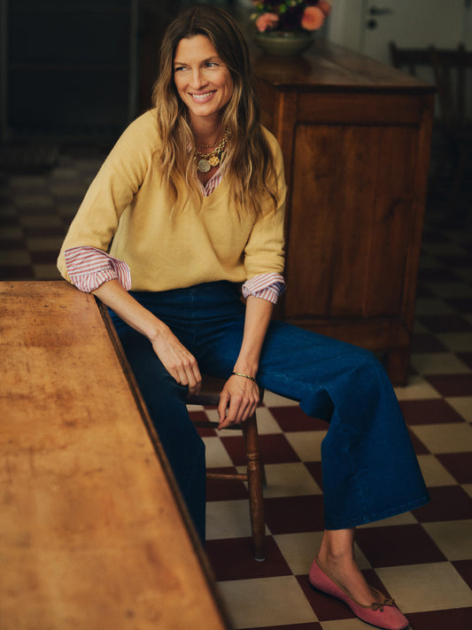 Woman wearing a Kerry Cashmere Sweater by J.McLaughlin layered over a striped blouse, paired with blue wide-leg jeans and pink flats, sitting indoors with a warm smile.