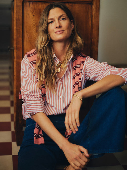 Woman sitting indoors wearing Finn Shirt in Stripe by J.McLaughlin, styled with layered gold jewelry and a striped sweater draped over her shoulders, embodying effortless style.