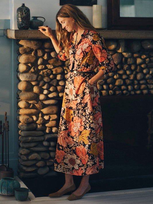 Woman wearing a Hannah Silk Blend Dress in City Garden by J.McLaughlin featuring a vibrant floral pattern, standing and smiling near a rustic stone fireplace, showcasing effortlessly stylish, enduring design.