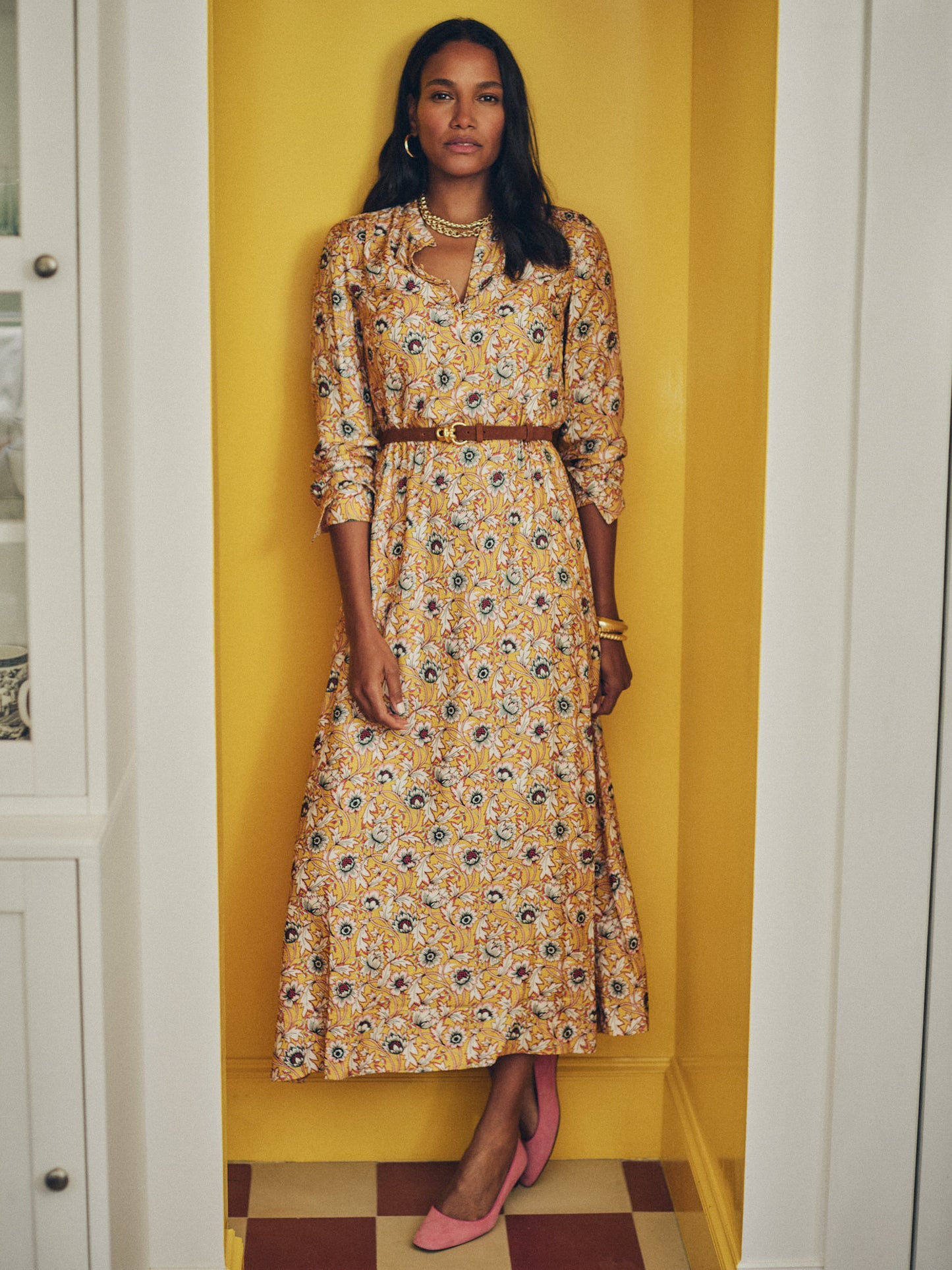 Woman wearing a Hannah Silk Blend Dress in Belle Epoque Bloom by J.McLaughlin with a delicate floral pattern, styled with a brown belt and pink shoes, standing confidently against a yellow wall.