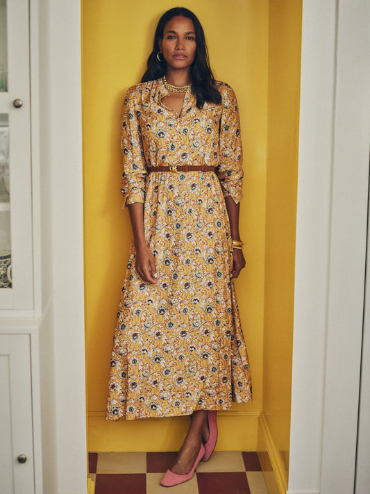 Woman wearing a Hannah Silk Blend Dress in Belle Epoque Bloom by J.McLaughlin with a delicate floral pattern, styled with a brown belt and pink shoes, standing confidently against a yellow wall.