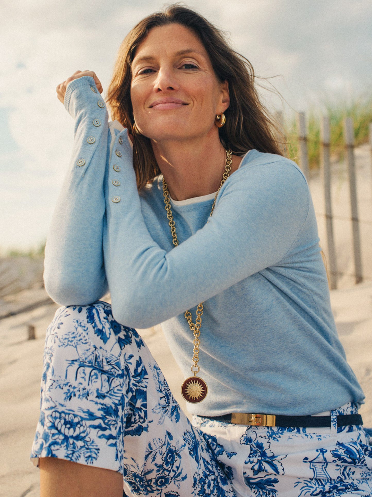 Woman wearing a light blue Jamey Sweater by J.McLaughlin with button details on the sleeve, paired with blue and white floral pants, a gold pendant necklace, and hoop earrings, sitting outdoors on a sandy beach.