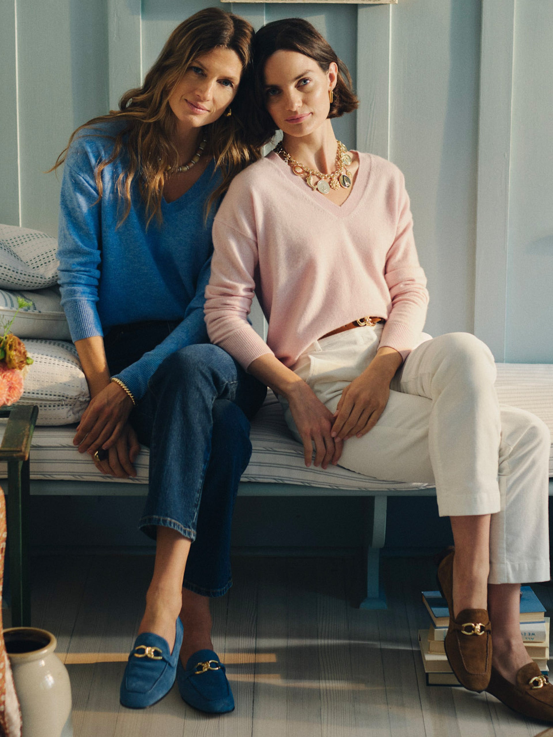 Two women seated indoors wearing stylish sweaters, one in a Kerry Cashmere Sweater from J.McLaughlin paired with white pants and a statement necklace, embodying effortlessly stylish, enduring designs.