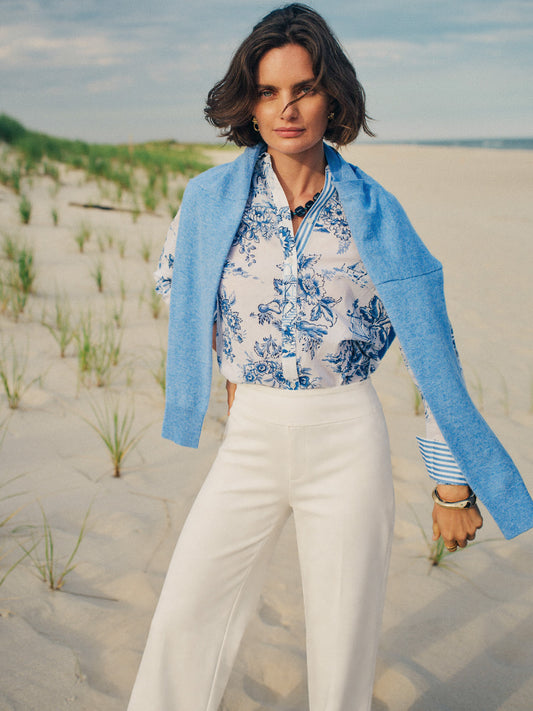 Woman stands on a beach wearing a Lois Shirt in Antique Toile Stripe by J.McLaughlin with a light blue sweater draped over her shoulders and white high-waisted pants.