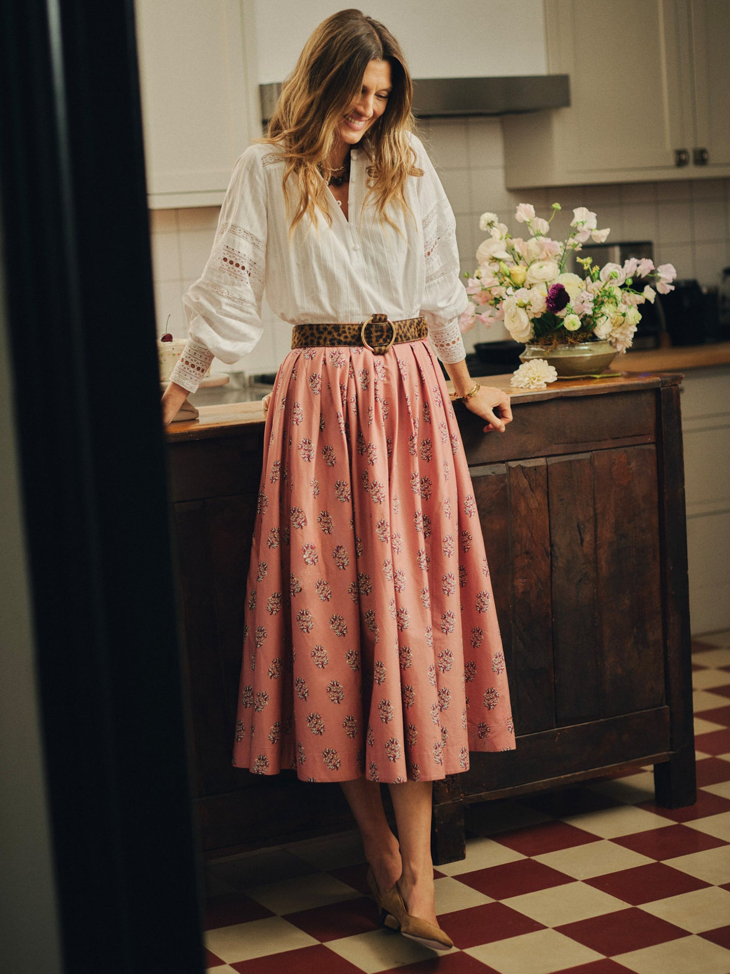 Woman wearing an Arabella Skirt in Heritage Bloom Cross by J.McLaughlin, paired with a white blouse and leopard print belt, standing in a classic kitchen with floral decor.