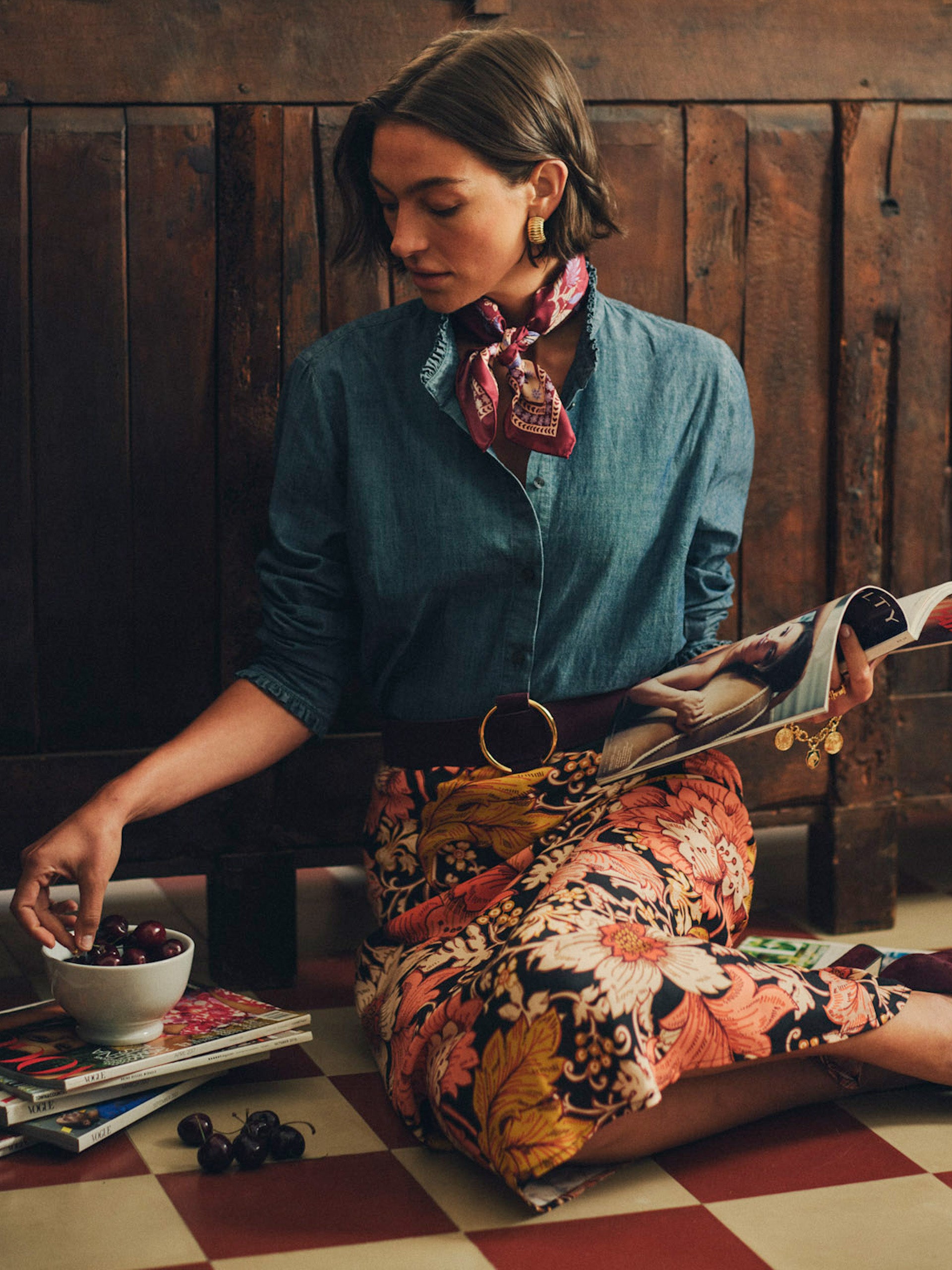 Woman wearing a Tess Ruffle Collar Chambray Shirt by J.McLaughlin styled with a floral skirt and red patterned scarf, sitting on a checkered floor reading a magazine and reaching for cherries.