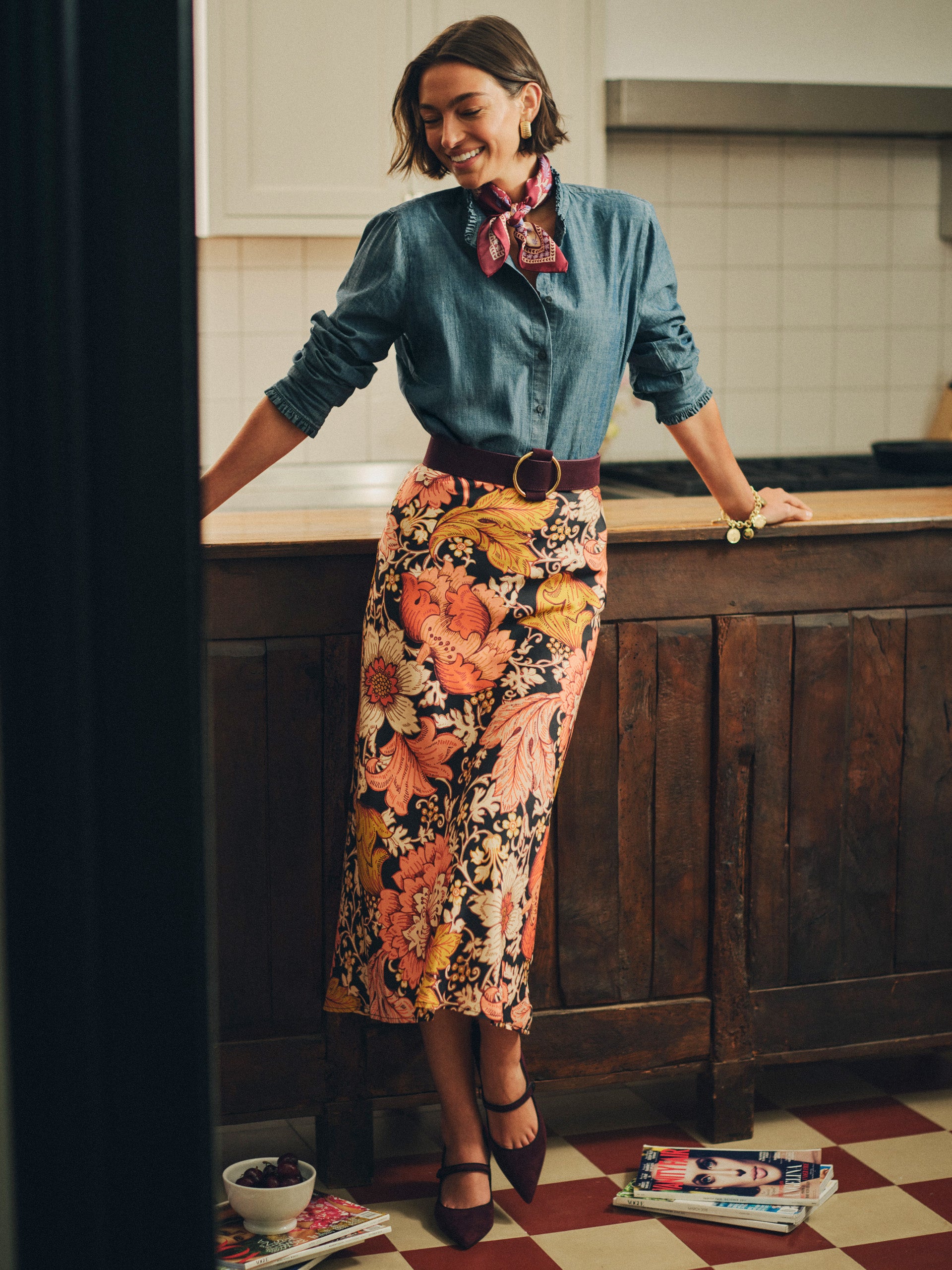 Woman leaning on kitchen counter wearing Adelyn Silk Blend Skirt in City Garden Bias with floral pattern, denim shirt, burgundy belt, and matching shoes by J.McLaughlin