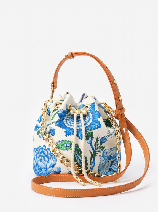 Daisy Bucket Bag in Mini Cerulean Garden by J.McLaughlin features a floral print in blue and green with tan leather straps, showcasing effortlessly stylish, enduring designs and a legacy of timeless fashion.