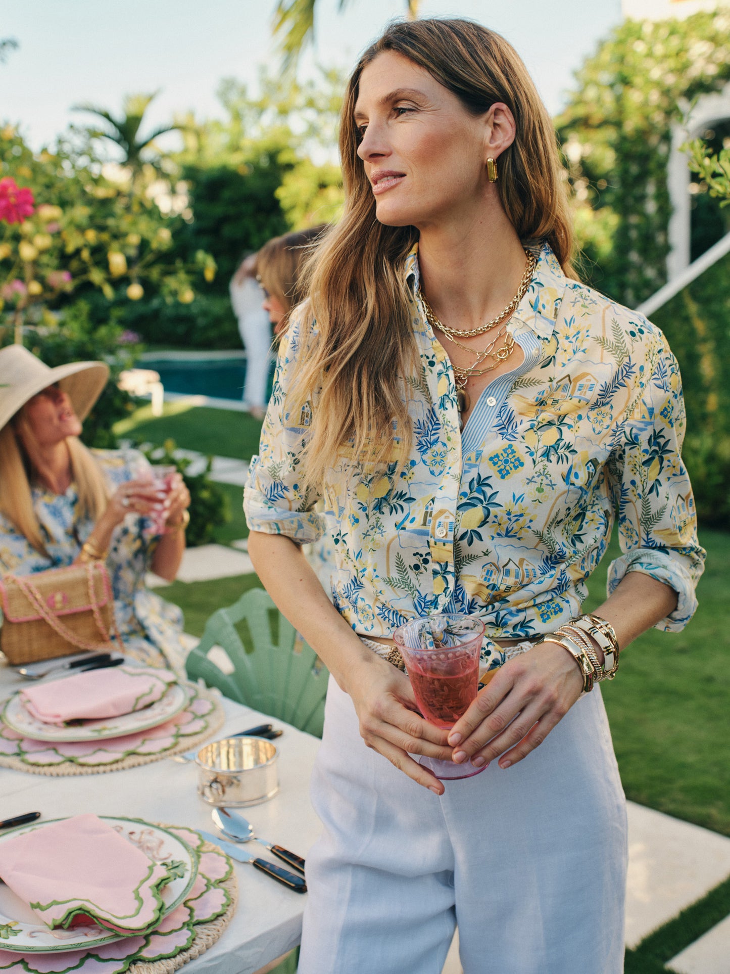 Woman wearing a Lois Shirt in Mini Amalfi Coast Stripe by J.McLaughlin, styled with white pants and gold accessories at an outdoor garden gathering, embodying effortlessly stylish, enduring designs and a legacy of timeless fashion. A destination for defining style. Life, Well-Lived.