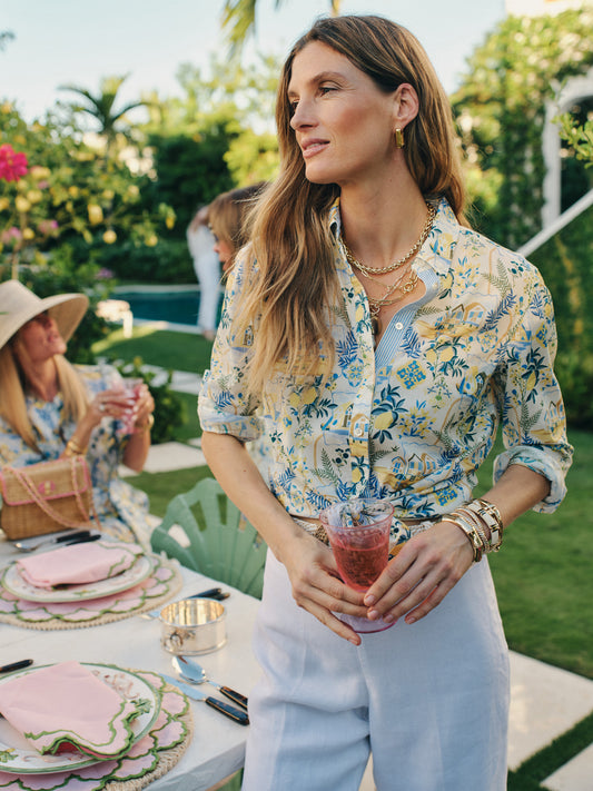 Woman wearing a Lois Shirt in Mini Amalfi Coast Stripe by J.McLaughlin, styled with white pants and gold accessories at an outdoor garden gathering, embodying effortlessly stylish, enduring designs and a legacy of timeless fashion. A destination for defining style. Life, Well-Lived.
