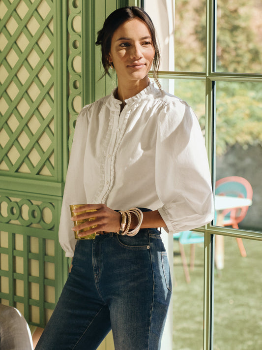 A woman wearing a white Josette Blouse by J.McLaughlin with puffed sleeves and delicate detailing, paired with dark jeans, standing by a sunlit window holding a glass, showcasing effortlessly stylish, enduring designs rooted in a legacy of timeless fashion. A destination for defining style. Life, Well-Lived.
