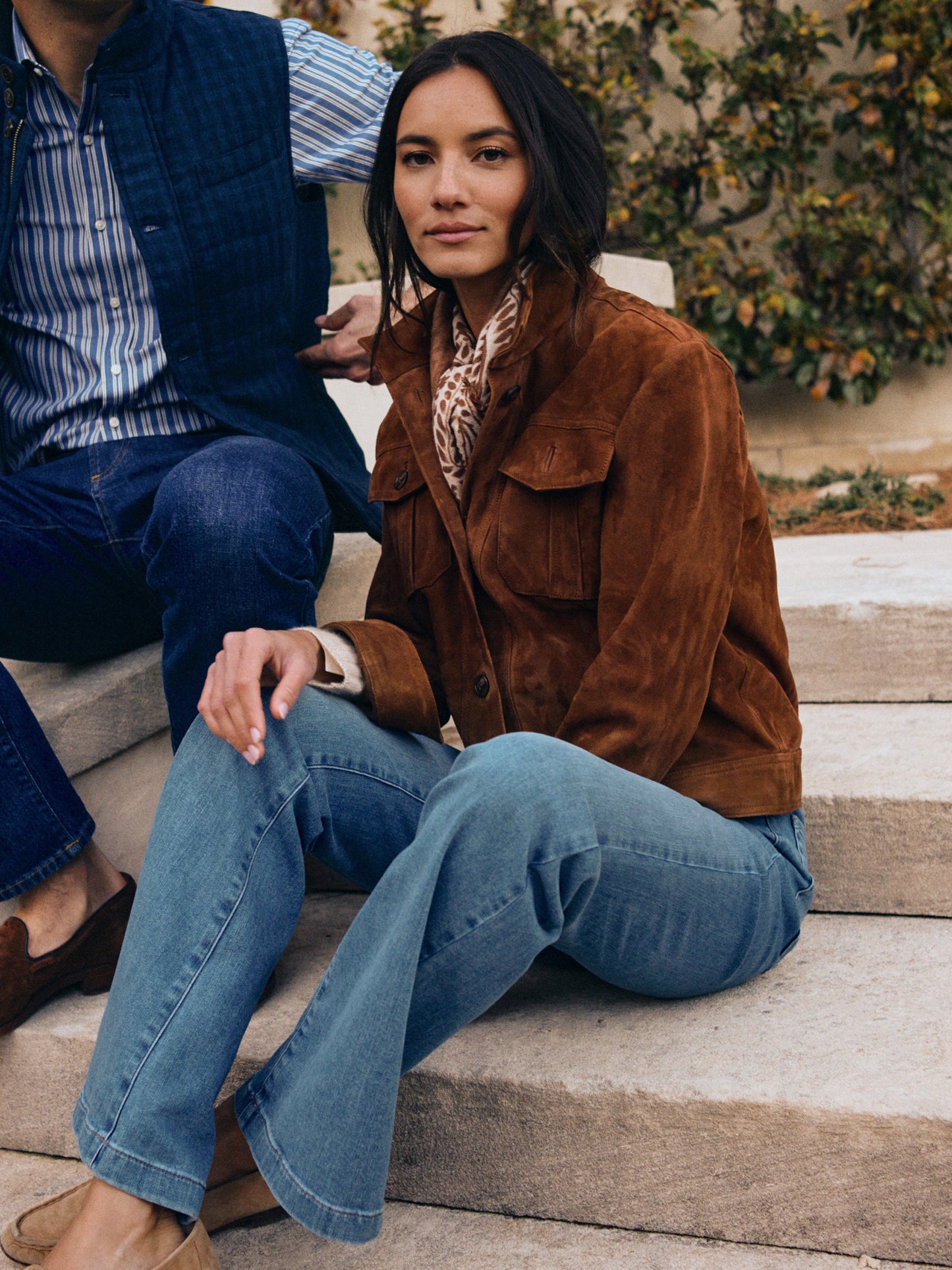 Woman wearing a brown Frances Suede Jacket by J.McLaughlin paired with blue jeans, sitting on stone steps beside a man in casual attire, showcasing effortlessly stylish, enduring designs.